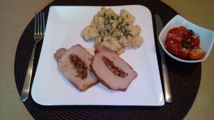 Ein gef&uuml;llter Schweiner&uuml;cken liegt auf einem wei&szlig;en Teller, umgeben von Kartoffeln und einer roten Sauce in einer kleinen Sch&uuml;ssel.