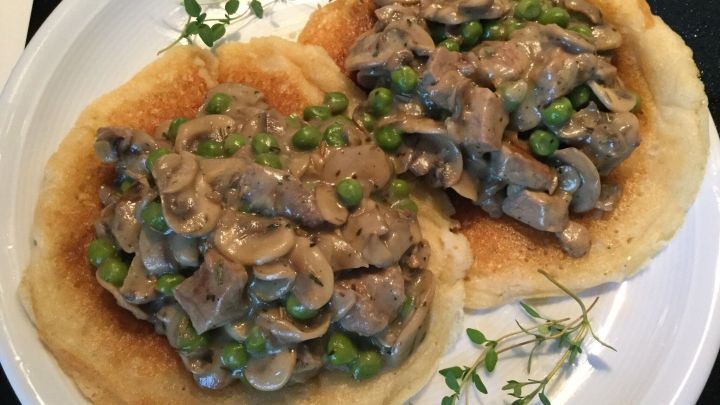 Zwei Blini befinden sich auf einem Teller, belegt mit einem Ragout aus Pilzen und Rinderfilet, umgeben von Kr&auml;utern.