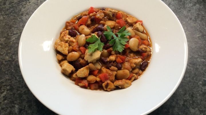 Ein Teller Chili con Carne mit H&uuml;hnerfleisch, gef&uuml;llt mit bunten Bohnen und frischen Kr&auml;utern auf einem dunklen Tisch.