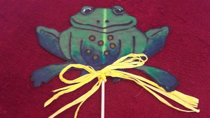 Ein bunter Pflanzenstecker in Form eines Frosches mit einem gelben Band ziert einen roten Untergrund.