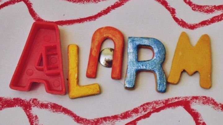 Bunte Buchstaben formen das Wort "ALARM" vor einem wellenf&ouml;rmigen, rot-wei&szlig; gestreiften Hintergrund, der Dringlichkeit signalisiert.