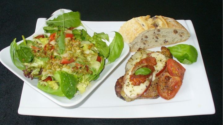 &Uuml;berbackenes Schweiner&uuml;ckensteak mit Mozzarella und Tomaten liegt auf einem wei&szlig;en Teller, begleitet von frischem Salat und Brot.