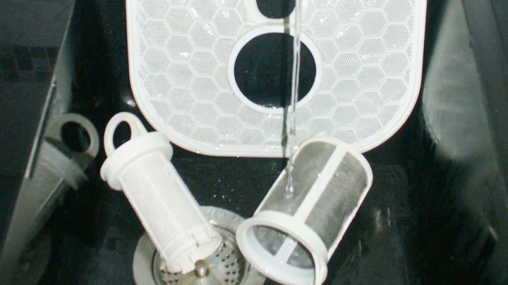 Eine wei&szlig;e Geschirrsp&uuml;ler-Schelle liegt in einer Sp&uuml;le, w&auml;hrend Wasser dar&uuml;ber flie&szlig;t; daneben befinden sich weitere Reinigungsutensilien.