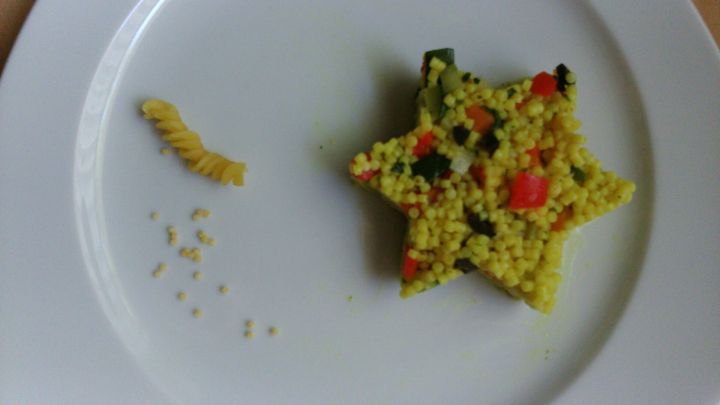 Ein bunter Graupennudelsalat hat die Form eines Sterns und liegt auf einem wei&szlig;en Teller, neben einer kleinen Pasta.