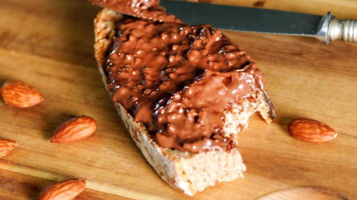 Ein St&uuml;ck Brot ist mit Nusscreme bestrichen und auf einem Holzbrett platziert, umgeben von einigen Mandeln.