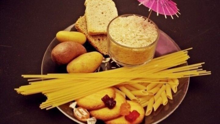 Ein Teller mit verschiedenen Lebensmitteln wie Pasta, Brot, Kartoffeln und Keksen, die ungesund wirken, steht vor einem dunklen Hintergrund.