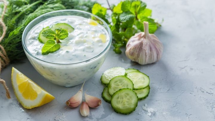 Ein cremiger Knoblauchdip in einer Sch&uuml;ssel umgeben von Gurkenscheiben und frischen Kr&auml;utern auf einem hellen Tisch.