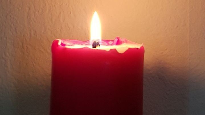 Eine rote Kerze leuchtet hell auf einem Tisch, w&auml;hrend im Hintergrund ein Bild von kahlen B&auml;umen an der Wand h&auml;ngt.