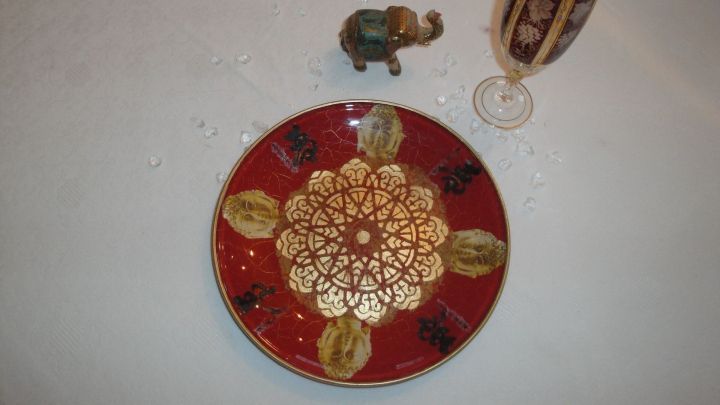 Ein dekorierter Glasteller mit goldenen Mustern liegt auf einem wei&szlig;en Tisch, umgeben von einem kleinen Elefanten und einem Glas.