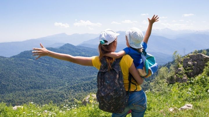 Eine Mutter h&auml;lt ein Kind auf dem R&uuml;cken und genie&szlig;t die Aussicht auf eine gr&uuml;ne Berglandschaft beim Reisen.
