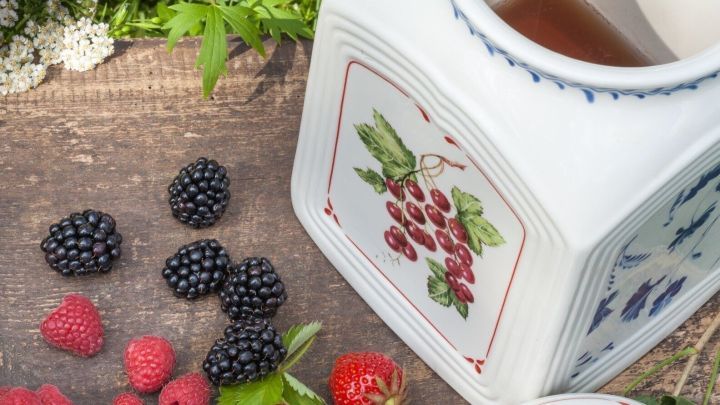 Der Rumtopf steht offen auf einem Holztisch, umgeben von frischen Beeren wie Himbeeren, Erdbeeren und Brombeeren.