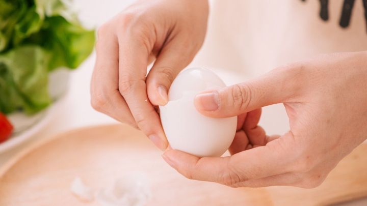 Eine Hand h&auml;lt ein gekochtes Ei und sch&auml;lt es vorsichtig, w&auml;hrend im Hintergrund frisches Gem&uuml;se sichtbar ist.