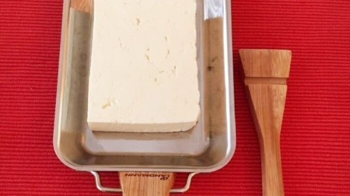 Eine Grillpfanne aus Edelstahl mit Feta und einem Holzpinsel liegt auf einem roten Untergrund, bereit zum Grillen.