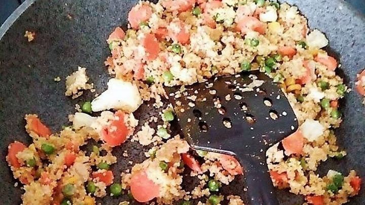 Ein bunter Reste-Couscous wird in einer Pfanne mit einer schwarzen K&uuml;chenhilfe zubereitet, umgeben von einer modernen K&uuml;che.