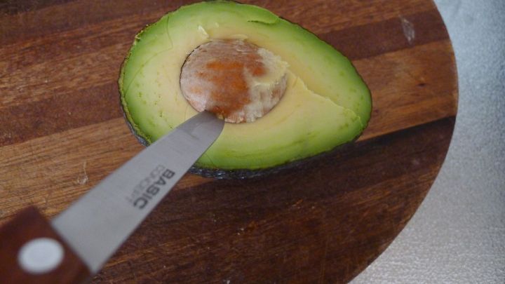 Eine halbierte Avocado liegt auf einem Holzbrett, w&auml;hrend ein Messer den Kern vorsichtig herausl&ouml;st.