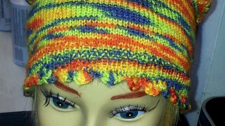 Eine bunte M&uuml;tze aus "Maschenmix" sitzt auf einem Kopf einer Puppe, umgeben von Pflegeprodukten auf einem Tisch.