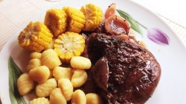 Eine geschmorte Entenkeule liegt auf einem Teller mit Gnocchi und Buttermais, umgeben von einem Glas Rotwein.