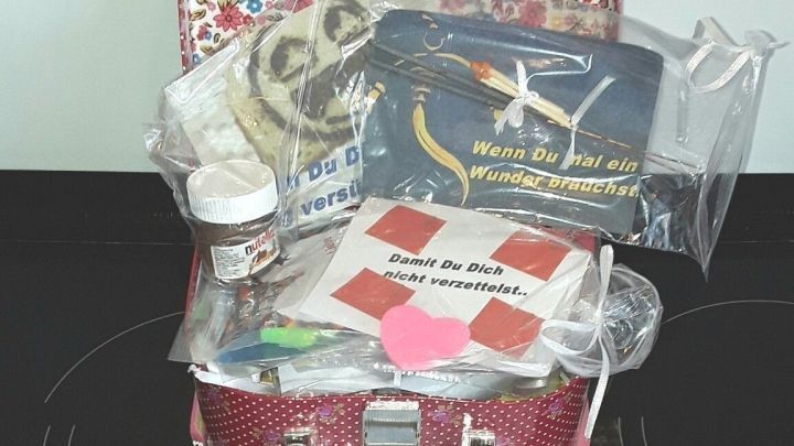 Ein bunter Koffer steht offen auf einer schwarzen Oberfl&auml;che, gef&uuml;llt mit verschiedenen Geschenken und buntem Papier.