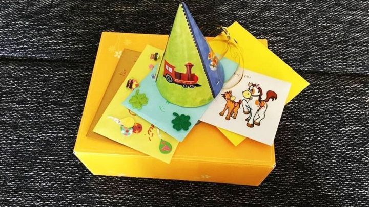 Ein gelbes Geschenkpaket mit einer bunten Geburtstagskarte und einer Partyh&uuml;te-Dekoration liegt auf einem dunklen Tisch.