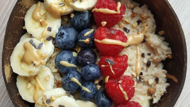 Eine Sch&uuml;ssel Porridge liegt auf einem hellen Holzuntergrund, garniert mit Bananen, Himbeeren und Blaubeeren.