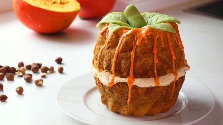 Ein Gugelhupf aus K&uuml;rbis, garniert mit Frischk&auml;se-Zimt-Creme, steht auf einem Teller neben einer K&uuml;rbisfrucht und &Auml;pfeln.