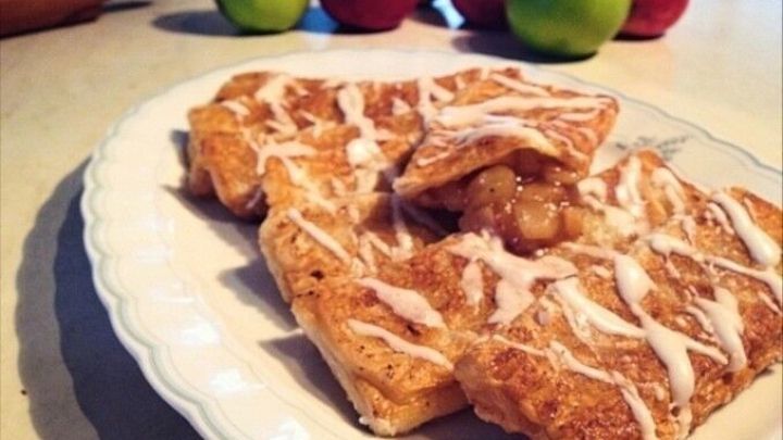 Apfeltaschen aus Bl&auml;tterteig liegen auf einem Teller, umgeben von frischen &Auml;pfeln und einer rollenden Teigrolle.