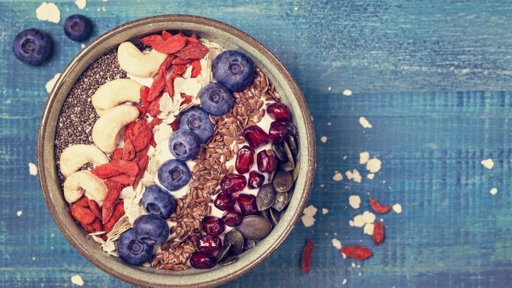 Eine Sch&uuml;ssel mit buntem Porridge, garniert mit Beeren und N&uuml;ssen, steht auf einer blauen Holzoberfl&auml;che.