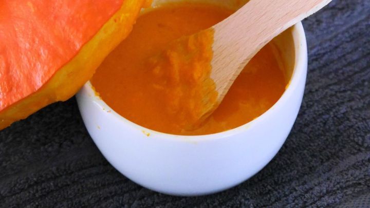 Eine K&uuml;rbis-Gesichtsmaske ist in einer wei&szlig;en Sch&uuml;ssel, w&auml;hrend der K&uuml;rbis und Dekorationskerzen im Hintergrund sichtbar sind.