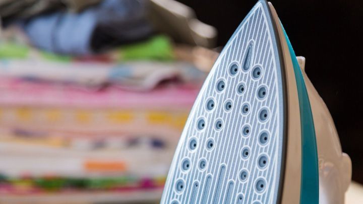 Die B&uuml;geleisensohle steht im Vordergrund und wirkt stark verschmutzt, w&auml;hrend gewaschene Kleidung im Hintergrund liegt.