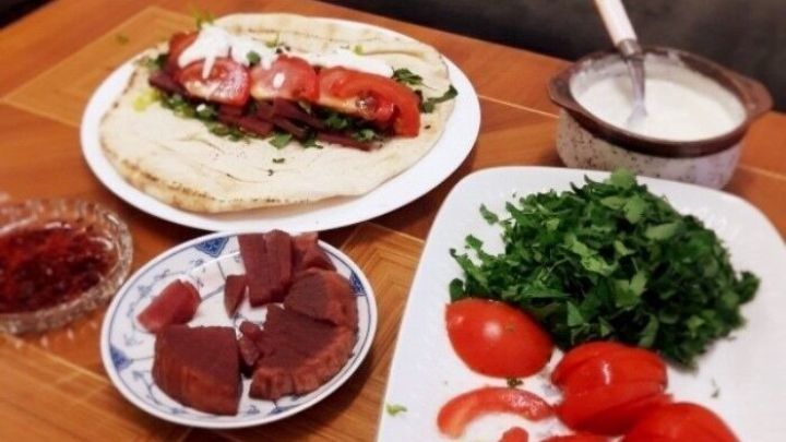 Auf einem Tisch liegt ein Falafel-Wrap, w&auml;hrend frische Tomaten, gr&uuml;ner Salat und So&szlig;en zur Zubereitung bereitstehen.