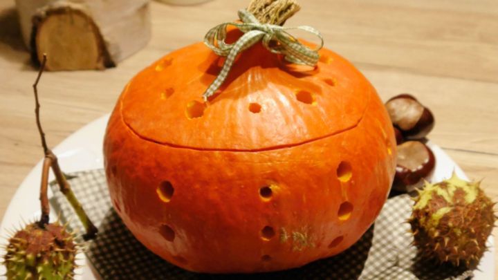 Ein orangefarbener K&uuml;rbis mit vielen L&ouml;chern steht auf einem wei&szlig;en Teller, umgeben von Maronen und Kastanien.