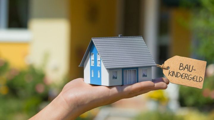 Eine Hand h&auml;lt ein kleines Modellhaus mit einem Schild "Baukindergeld" vor einem unscharfen, hellen Hintergrund.