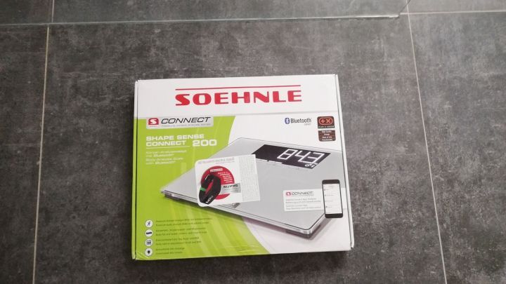 Eine digitale Personenwaage liegt auf einem grauen Fliesenboden, bereit zur Nutzung zur Unterst&uuml;tzung von Ess- und Trinkgewohnheiten.