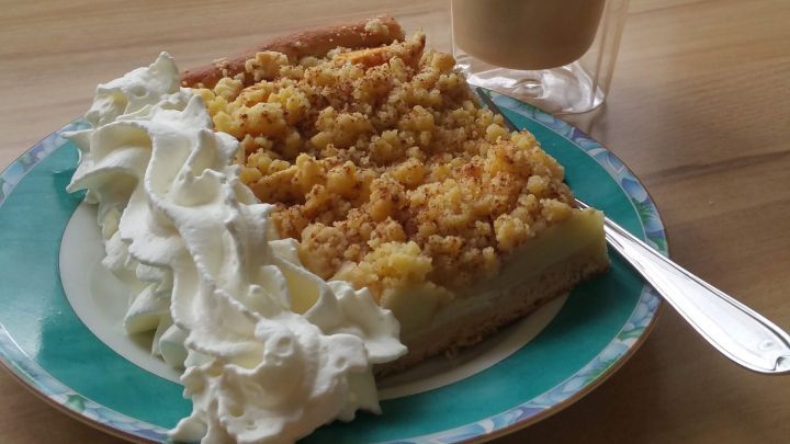 Ein saftiges St&uuml;ck Apfelkuchen mit Streuseln wird auf einem Teller mit Sahne serviert, daneben steht ein Glas Kaffee.