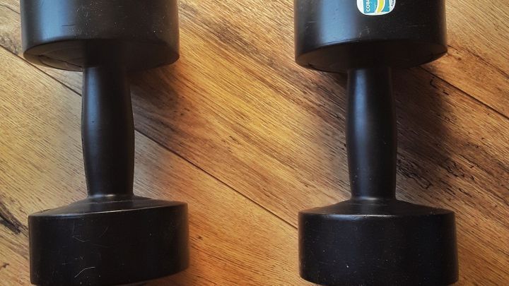Zwei schwarze Hanteln liegen auf einem hellen Holzfu&szlig;boden und warten darauf, f&uuml;r Bewegung und gesunde Ern&auml;hrung genutzt zu werden.