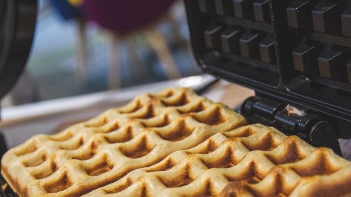 Ein Waffeleisen &ouml;ffnet sich, w&auml;hrend frisch gebackene Waffeln auf der Unterseite liegen, umgeben von einem gem&uuml;tlichen Ambiente.