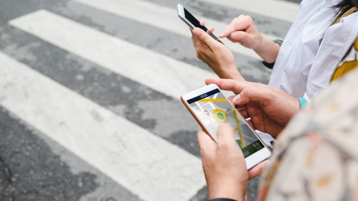 Zwei Personen stehen an einem Zebrastreifen und nutzen ihre Smartphones zur Navigation in einer st&auml;dtischen Umgebung.