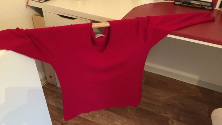 Ein roter Kaschmirpullover h&auml;ngt auf einem Kleiderb&uuml;gel, w&auml;hrend er in einem hellen Raum auf einem Tisch trocknet.