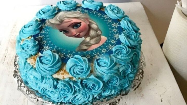 Ein blauer Kuchen mit Marschmallow-Creme-Topping, verziert mit Rosen und einem Bild von einer Figur, steht auf einem wei&szlig;en Tisch.