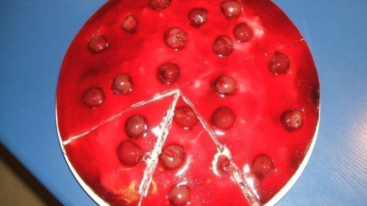 Eine runde, rote Torte mit Kirsch-Topping, teilweise angeschnitten, auf einem blauen Tisch pr&auml;sentiert.