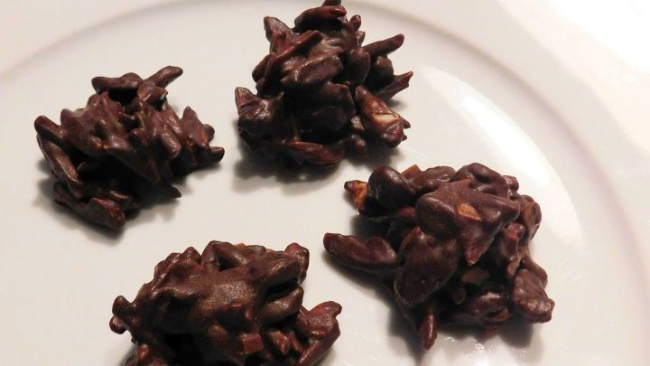 Dunkle Schoko-Mandel-Knusperh&auml;ufchen liegen auf einem wei&szlig;en Teller, umgeben von Schokolade und Mandelst&uuml;cken.