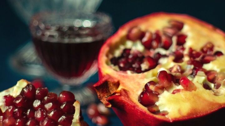 Ein halbierter Granatapfel liegt auf einem Tisch, aus dem rote Kerne und Saft flie&szlig;en, umgeben von einem Glas Saft.