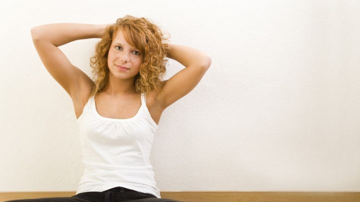 Eine Frau mit Naturlocken sitzt entspannt am Boden und streicht sich den Haaren zur&uuml;ck, vor einer hellen Wand.