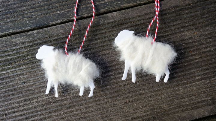 Zwei Weihnachts-Schafe aus wei&szlig;en Materialien h&auml;ngen an einer roten Schnur und sind auf einer dunklen Holzoberfl&auml;che platziert.