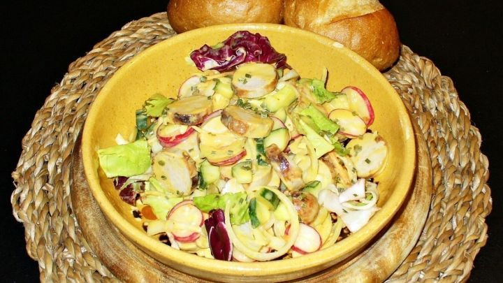 Ein bunter Bratwurstsalat mit frischem Gem&uuml;se steht in einer Sch&uuml;ssel, umgeben von zwei Br&ouml;tchen auf einem Teller.