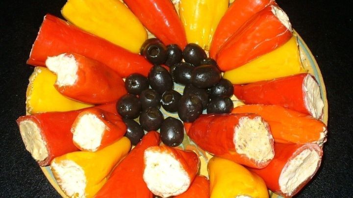 Bunte, s&uuml;&szlig;saure Spitzpaprika sind auf einem Teller angeordnet, gef&uuml;llt mit verschiedenen K&auml;sesorten und umgeben von Oliven.