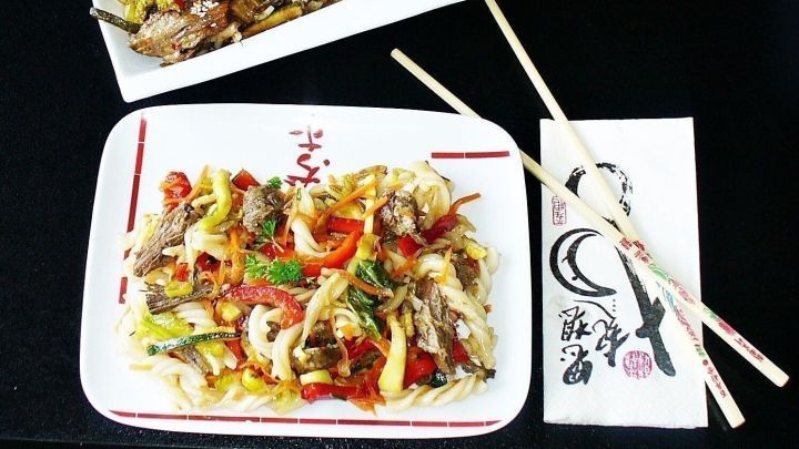 Ein asiatischer Rindfleischsalat liegt in einer wei&szlig;en Schale neben einer Rindfleischpfanne auf einem dunklen Tisch.