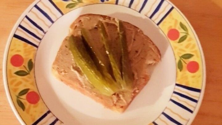 Ein St&uuml;ck Brot mit einem w&uuml;rzigen Brotaufstrich und Gurkenscheiben liegt auf einem bunten Teller auf einem Holztisch.