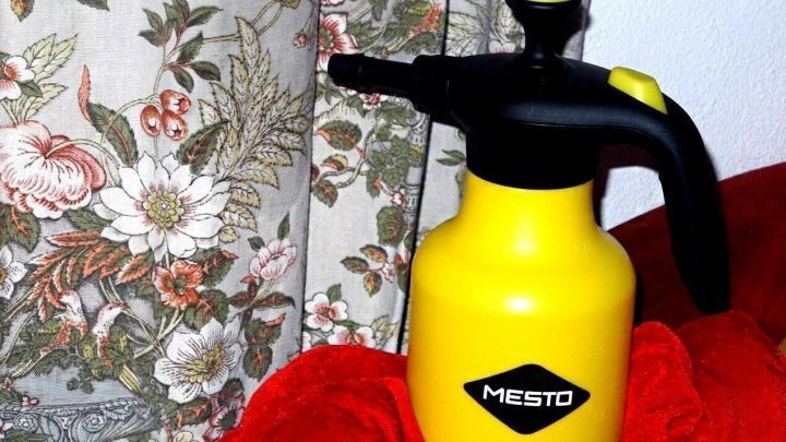 Ein gelbes Spr&uuml;hger&auml;t steht auf einem roten Tuch vor einem blumigen Vorhang, bereit, die Luftfeuchtigkeit in Wohnr&auml;umen zu erh&ouml;hen.