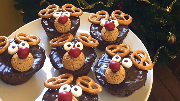 Die fluffigen Rentiermuffins auf einem Teller, dekoriert mit Brezeln und roter Nase, stehen festlich vor einem Weihnachtsbaum.